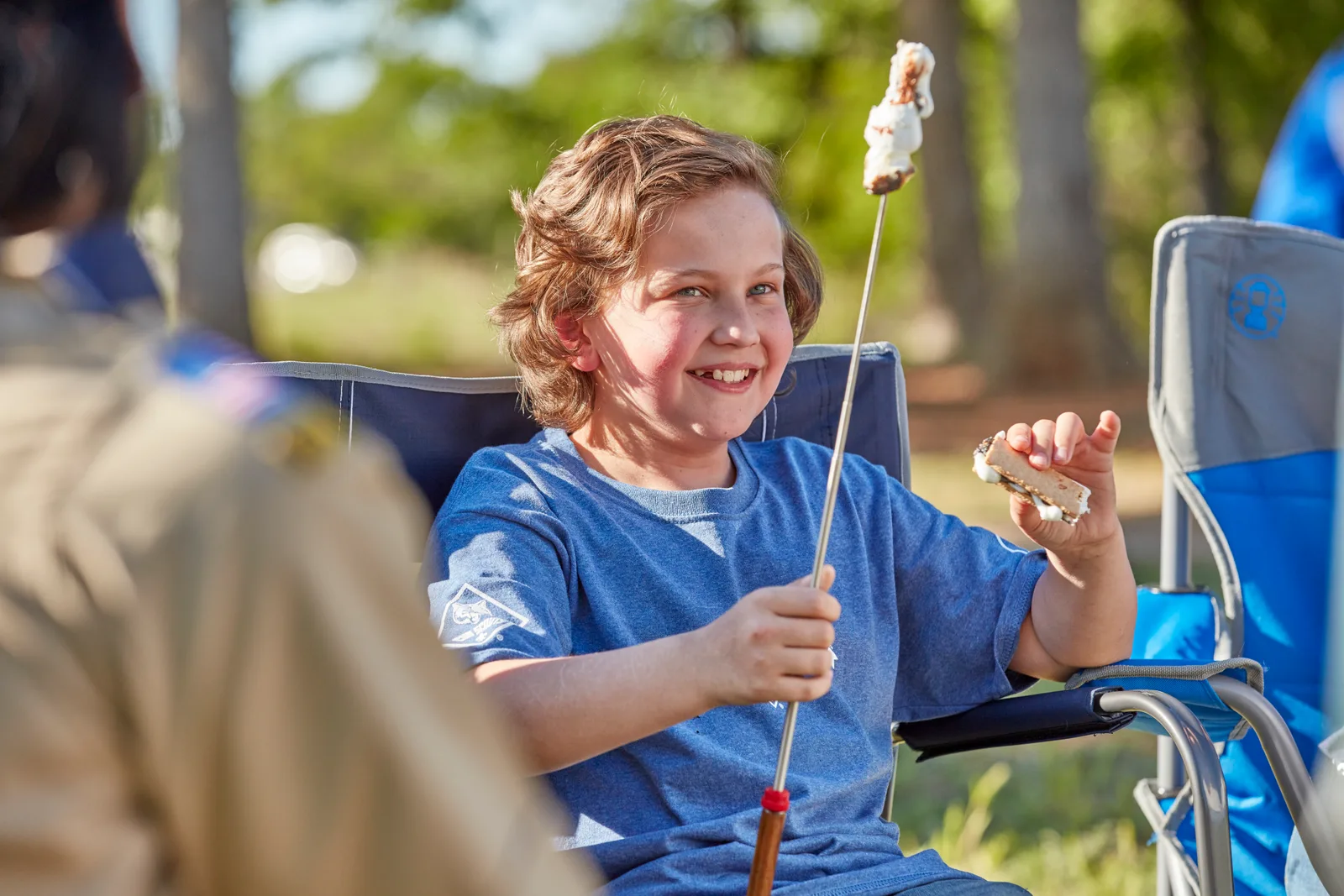 A Cub Scout grinning as he holds up a toasted marshmallow on a stick