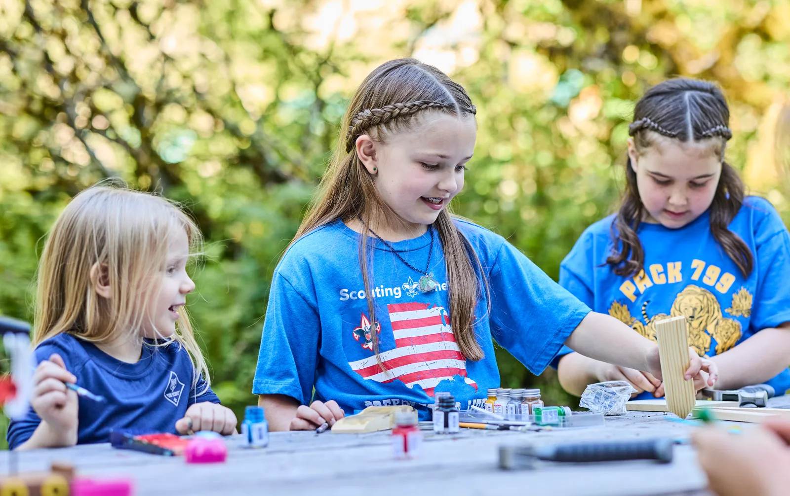 Three Cub Scouts sanding and painting Pinewood Derby cars at an outdoor table