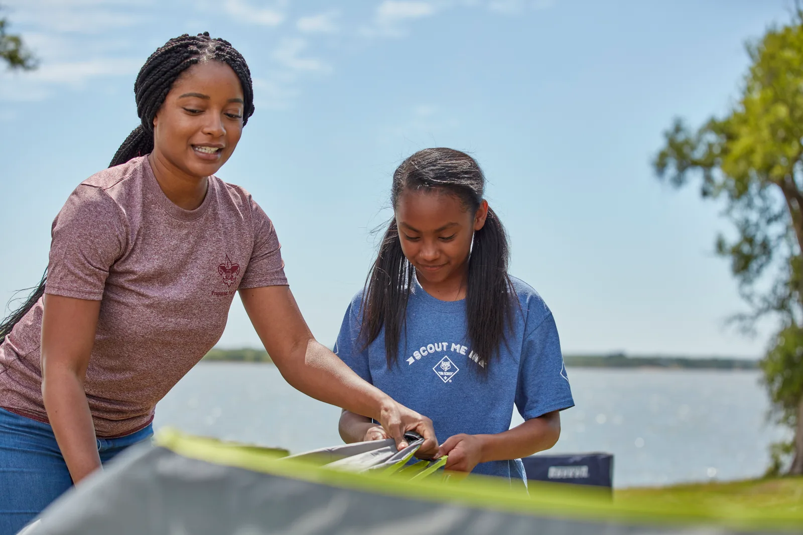 A parent and a Cub Scout threading poles through a tent beside a lake